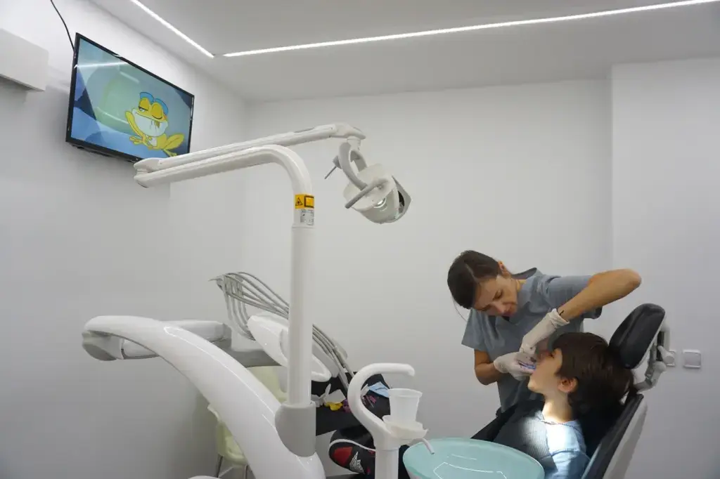 Dentista revisant la boca d’un nen assegut a la consulta, mentre el nen mira atentament.