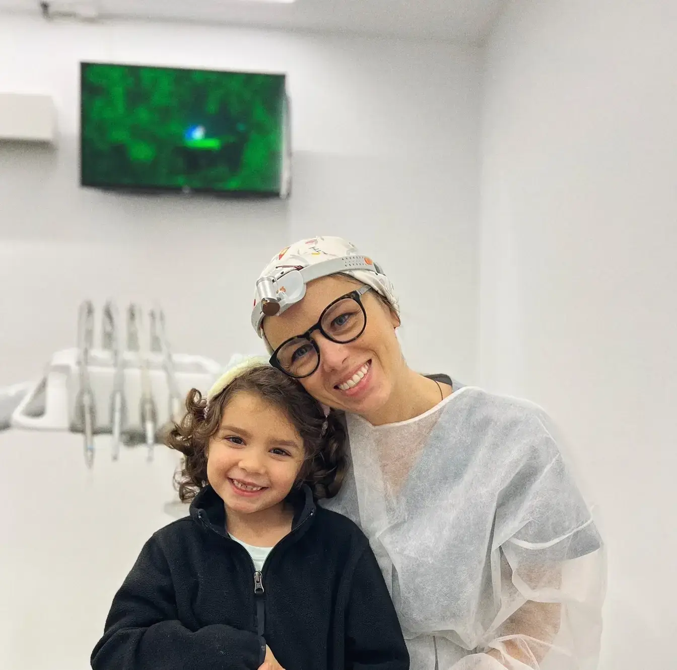 Dentista infantil i nena petita somrient juntes a la consulta, transmetent proximitat i confiança durant la revisió dental.